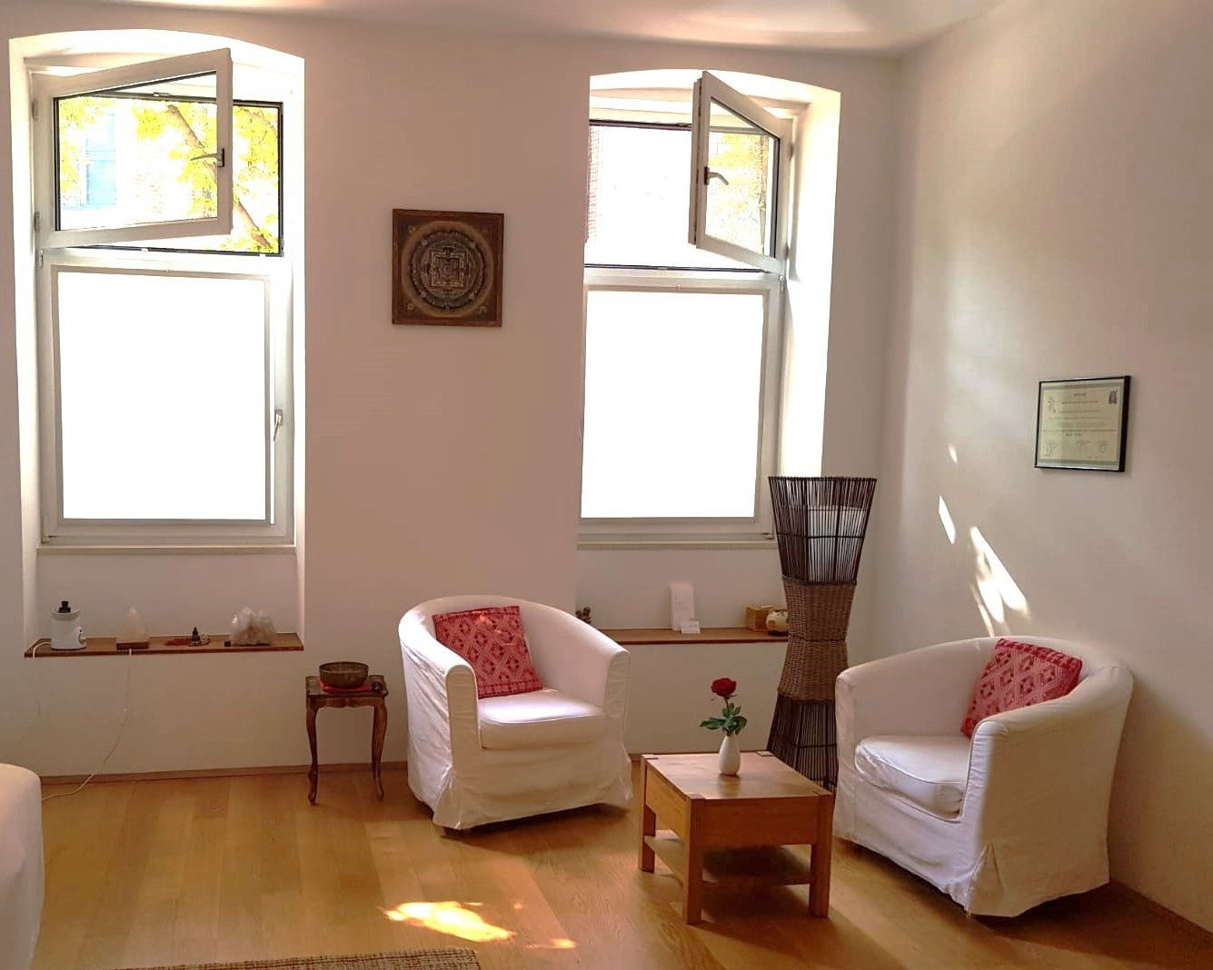Freundlich heller Raum mit zwei weißen Couch-Sesseln, einem kleinen Holztisch mit einer Vase, zwei offenen Fenstern und einer Stehlampe aus Rattern auf Holzboden.